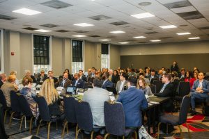A large group of people seated around tables in a conference room, engaged in discussion. Some attendees are taking notes or looking at laptops, while others are listening attentively. The room has large windows and a modern design