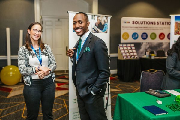 A woman with glasses and a black and white patterned sweater stands next to a man in a suit with a green pocket square. They are smiling and posing at a conference or event, with banners and tables in the background displaying various informational materials