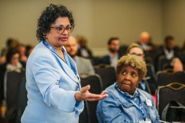 A woman with curly black hair and glasses speaks to an audience, gesturing with her hand. She is wearing a light blue blazer. In the foreground, another woman with curly blonde hair listens attentively. The setting appears to be a conference or meeting room with attendees seated in the background