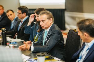 A group of professionals is seated around a conference table, engaged in discussion. One man in a suit is speaking, gesturing with his hands. Others are listening attentively, with some taking notes. The setting appears to be a business meeting or conference