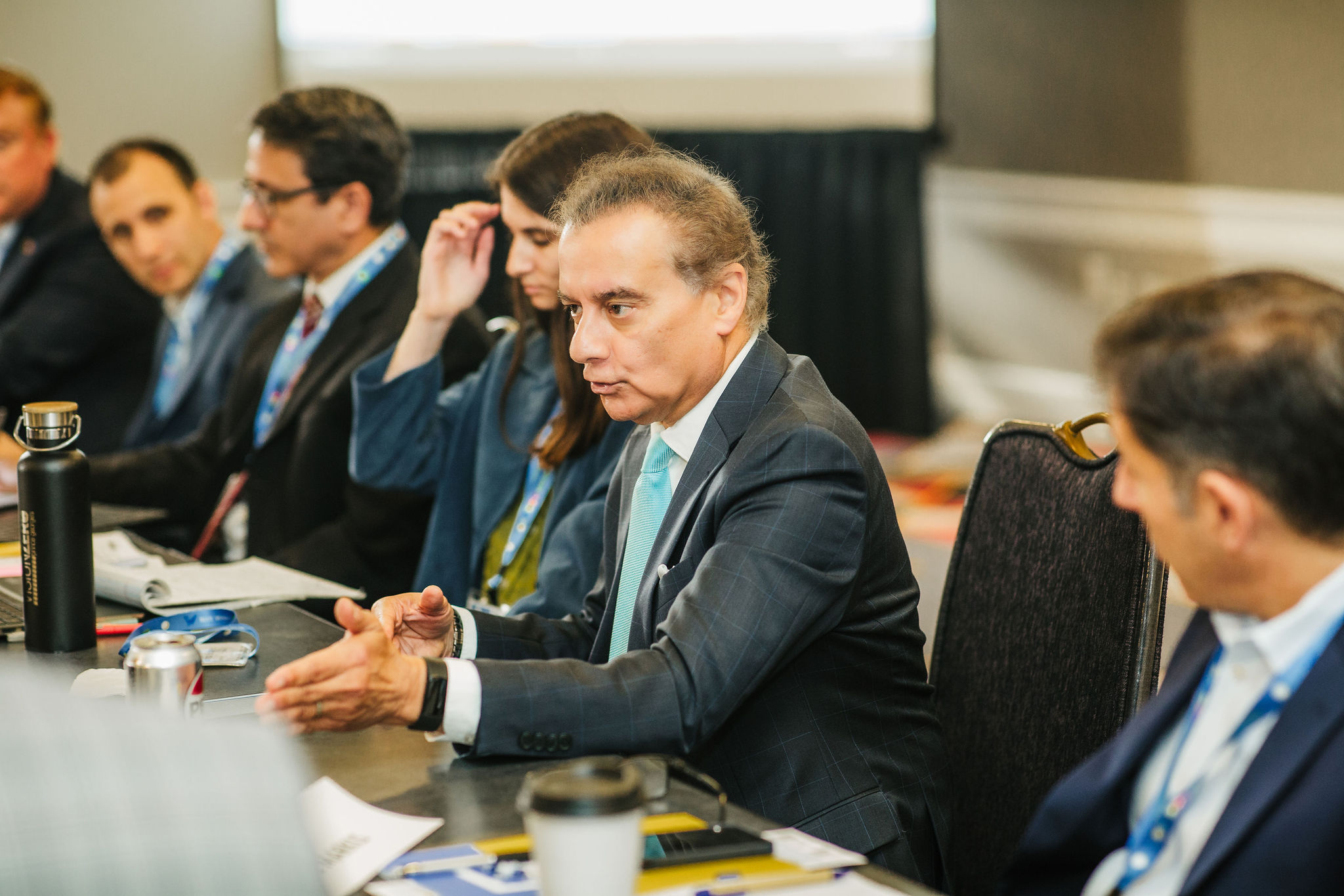 A group of professionals is seated around a conference table, engaged in discussion. One man in a suit is speaking, gesturing with his hands. Others are listening attentively, with some taking notes. The setting appears to be a business meeting or conference