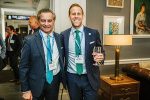 Two men stand together in a well-lit indoor setting. One man, wearing a dark suit and a blue tie, smiles and holds a glass. The other man, dressed in a lighter suit with a green tie, also smiles.