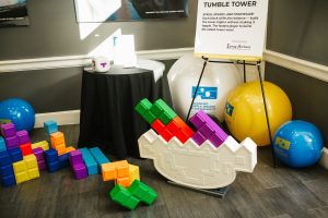 A colorful Tetris-inspired stacking game is displayed on a table, featuring various geometric blocks in bright colors like purple, orange, green, and blue. Nearby, a sign titled "Tumble Tower" explains the game, encouraging players to stack and balance the blocks. Large inflatable balls in blue and yellow are also present in the background
