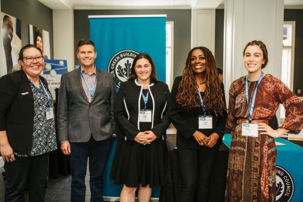 A group of five professionals stands together in a conference setting. They are smiling and wearing name tags. The backdrop features a banner with a logo related to green building. The individuals are dressed in business attire, with a mix of styles including a suit, a dress, and casual wear