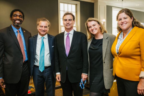 Five individuals stand together in a well-lit indoor setting. They are dressed in professional attire, with two men in suits and ties, and three women in business outfits. The group appears to be engaged in a formal or networking event