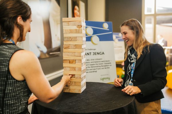 Two women are engaged in a game of Jenga at a table. One woman is pulling a wooden block from the tower, while the other watches with a smile. In the background, there is a sign with information about the event. The setting appears to be a lively indoor space