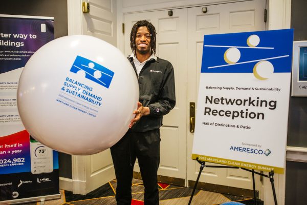 A person stands holding a large white balloon with a blue logo, next to a sign that reads "Networking Reception" and includes details about the event. The setting appears to be a conference or networking event, with a formal backdrop