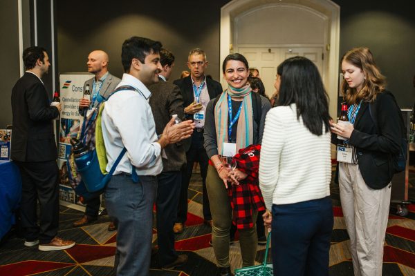 A group of professionals engages in conversation at a networking event. Two individuals in the foreground are smiling and talking, while others are gathered in the background, some holding drinks. The setting features a conference room with a banner visible on the left side
