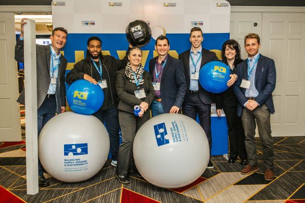 A group of seven people stands together in a conference setting, smiling and posing for the camera. They hold large blue and gray inflatable balls with logos. The background features a banner with the text "Building Business Connections