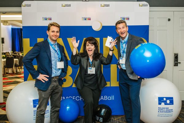 Three people are posing for a photo at an event. The woman in the center is smiling widely and holding a phone, while the two men on either side are making playful gestures. They are surrounded by large blue and white balloons, and a backdrop with event branding is visible behind them