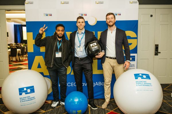 Three men stand together in front of a large blue and white backdrop with the text "HAVING ABLE." Two men are smiling and posing for the camera, one making a peace sign. The third man is holding a black helmet. Large blue and white balls are positioned in the foreground