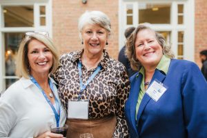 Three women are standing together, smiling at the camera. They are at a social event, with a blurred background of other attendees.