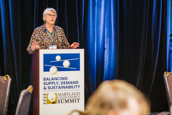 A woman with short gray hair, wearing a leopard print blouse, stands at a podium speaking. The podium displays a sign for the Maryland Clean Energy Summit, with the theme "Balancing Supply, Demand & Sustainability." In the background, there are blue curtains and blurred attendees seated at tables