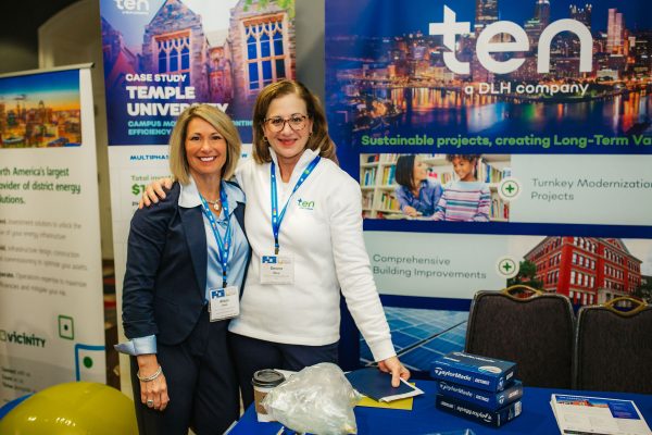 Two women stand together at a conference booth. They are smiling and posing in front of a backdrop featuring the company logo and information about sustainable projects.