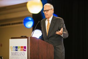 A man in a dark suit and glasses stands at a podium, speaking into a microphone. The podium has a sign that reads "Maryland Clean Energy Center." Colorful round decorations are visible in the background