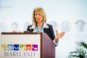 A woman with shoulder-length blonde hair speaks at a podium, gesturing with her hands. She is wearing a dark blazer over a light-colored blouse and has glasses resting on her head. The background features a presentation screen and a sign that reads "MARYLAND" with various colorful icons