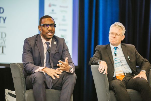 Two men are seated on stage during a panel discussion. One man, wearing glasses and a dark suit, is speaking into a microphone, gesturing with his hands. The other man, dressed in a suit with a striped tie, is listening attentively. A backdrop with logos and text is visible behind them