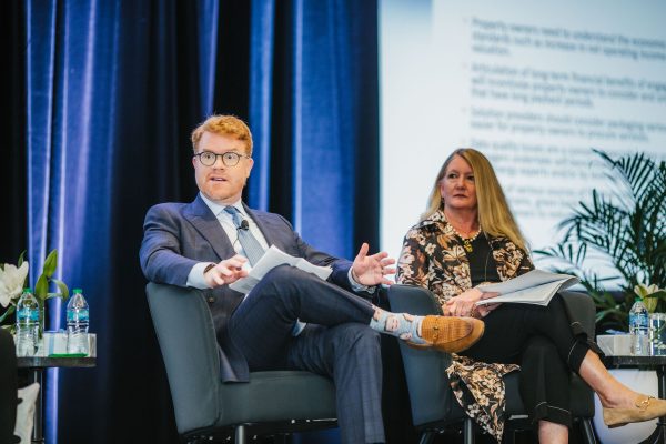 A man with red hair and glasses, wearing a blue suit and holding a notepad, is speaking while seated in a chair on stage. Next to him, a woman with long blonde hair, dressed in a black outfit with a floral print jacket.