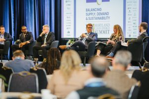 A panel discussion at the Maryland Clean Energy Summit featuring five speakers seated on a stage. The backdrop displays the event's title, "Balancing Supply, Demand & Sustainability." Attendees are visible in the foreground, listening to the discussion