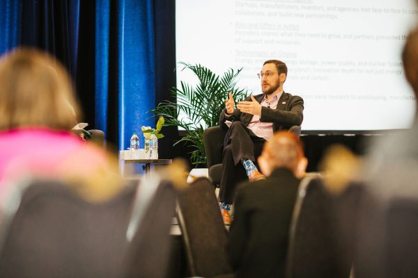 A man in a pink shirt and blazer sits on a stage, speaking to an audience. He gestures with his hands while discussing a topic, with a plant and water bottles visible on a table beside him. The audience is seated in the foreground, facing him