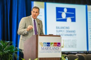A man in a suit stands at a podium with a sign that reads "MARYLAND CLEAN ENERGY CENTER." He is speaking into a microphone, with a presentation screen behind him displaying the text "Balancing Supply, Demand, Sustainability." The setting appears to be a conference or summit