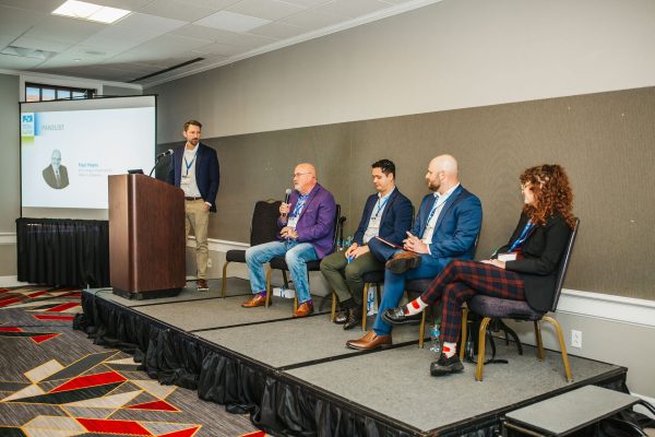 A panel discussion taking place in a conference room. Five individuals are seated on a stage, with one person standing at a podium. The seated panelists are engaged in conversation, while a presentation screen is visible in the background. The audience is not shown