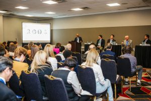 A panel discussion taking place in a conference room. Five speakers are seated at a table in front of a large screen displaying the number "2002." An audience of attendees is seated in rows, facing the speakers. The room has a professional setting with a mix of individuals engaged in the presentation
