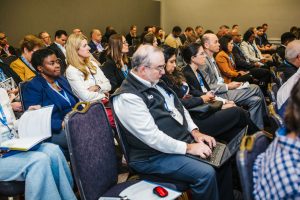 A diverse group of people seated in a conference room, attentively listening to a presentation. Some individuals are taking notes or using laptops, while others are engaged in discussion. The audience includes men and women of various ages and backgrounds