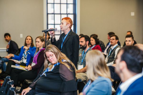 A person with short, bright red hair stands in a conference room, holding a microphone and asking a question. The audience, seated in rows, includes a diverse group of individuals engaged in the discussion, some looking towards the speaker