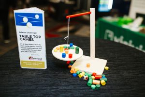A balance scale model is displayed on a table, featuring a circular platform with colorful cylindrical and disc-shaped pieces scattered around it. A sign next to the model reads "Balance Challenge: Table Top Games," explaining the activity's focus on balance and sustainability