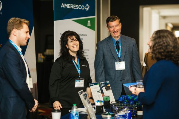 A group of four people engage in conversation at a conference booth. Two men and two women are present, with one woman smiling while speaking. The booth features a banner for Ameresco and displays promotional materials, including brochures and pens
