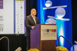 A man stands at a podium with a sign that reads "Maryland Clean Energy Center." He is speaking at an event, wearing a dark suit and a checkered shirt. The background features blue lighting and decorative elements