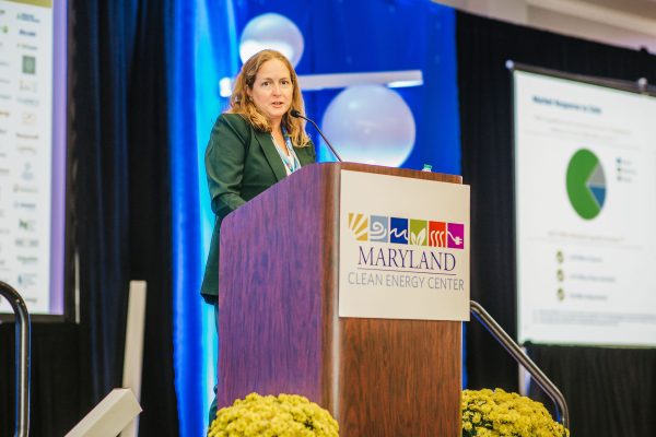 A woman with long brown hair speaks at a podium labeled "Maryland Clean Energy Center" during a conference. She is wearing a green blazer and appears to be presenting information to an audience, with a presentation screen visible in the background