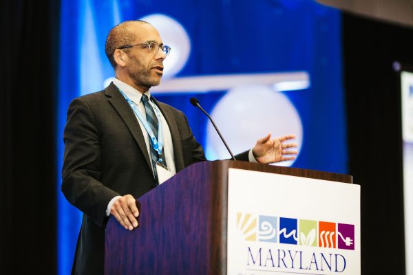 A man in a suit stands at a podium, speaking into a microphone. He has glasses and is gesturing with his right hand. The podium features a sign that reads "MARYLAND" along with various colorful symbols. A blue backdrop is visible behind him
