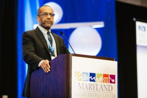 A man stands at a podium with a sign that reads "Maryland Clean Energy Center." He is wearing a suit and glasses, looking out at an audience. The background features blue and white decorations