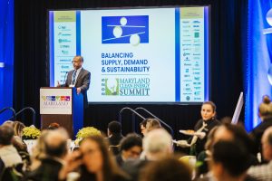 A speaker stands at a podium during the Maryland Clean Energy Summit, addressing an audience. Behind the speaker, a large screen displays the event title "Balancing Supply, Demand & Sustainability" along with various sponsor logos. Attendees are seated at tables, some engaged in conversation