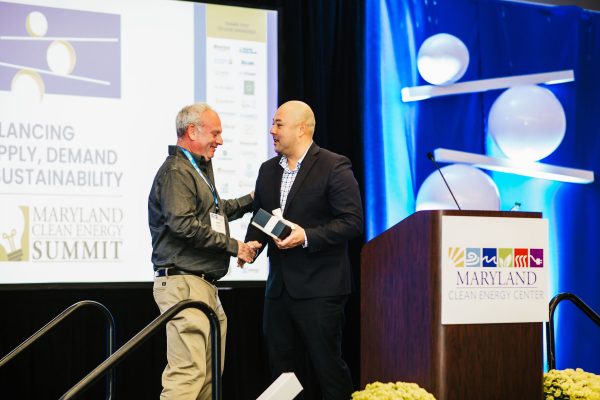 Two men are shaking hands on stage at the Maryland Clean Energy Summit. One man is handing a small award or gift to the other. In the background, a presentation screen displays information related to the event, and the stage is decorated with blue and white lighting