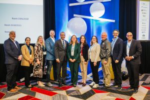 A group of ten people stands together in a conference setting, smiling at the camera. They are dressed in professional attire and are positioned in front of a backdrop featuring blue and white geometric shapes. The setting appears to be a business or educational event