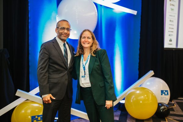 A man in a suit and a woman in a green suit stand together, smiling at the camera. They are in front of a blue and white backdrop with balloons and decorative elements. The setting appears to be a conference or event