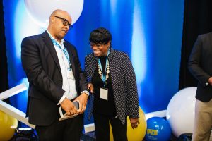 Two individuals are engaged in conversation at an event. One person, wearing glasses and a black suit, appears to be listening attentively, while the other, dressed in a polka-dot jacket, is smiling and gesturing. The background features blue lighting and large colorful balloons