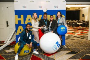 Five individuals pose together in a conference setting, smiling and holding large colorful balls. They are standing in front of a backdrop with the text "HAVING A BALL." The room features a carpet with geometric patterns and empty chairs in the background
