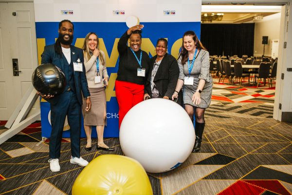 Five people stand in front of a large blue backdrop. The people are smiling and posing with oversized colorful balls.