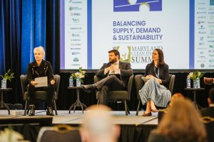 A panel discussion featuring three speakers seated on stage. The first speaker, a woman with short hair and a black jacket, holds a paper and is speaking. The second speaker, a man in a dark suit, listens attentively. The third speaker, a woman in a striped dress, also listens. A large screen in the background displays the title "Balancing Supply, Demand & Sustainability" along with the logo for the Maryland Clean Energy Summit. Small plants and water bottles are placed on the tables in front of the speakers