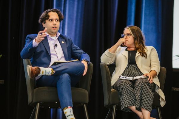 A man in a blue suit and colorful socks gestures while speaking at a panel discussion. Next to him, a woman in a light-colored cardigan sits with her arms crossed, listening attentively. Both are seated on stage with a dark backdrop