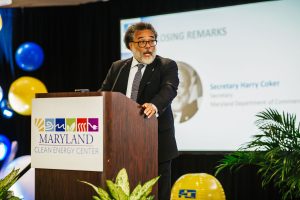 A man with curly hair and glasses stands at a podium, delivering remarks at an event. He is wearing a suit and tie, and there are colorful balloons in the background. A screen behind him displays text related to the Maryland Clean Energy Center