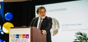 A man with curly hair and glasses stands at a podium, delivering remarks at an event. He is wearing a suit and tie, and there are colorful balloons in the background. A screen behind him displays text related to the Maryland Clean Energy Center