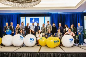 A diverse group of people stands and sits on a stage in front of a large screen displaying a logo. They are posing with large beach balls in various colors.