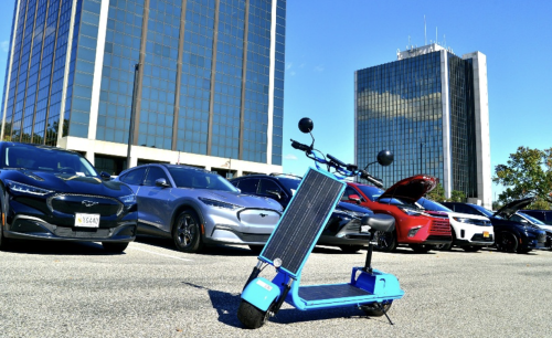 A blue electric scooter with a solar panel on the front is parked in a lot filled with various cars, including several sedans and SUVs. In the background, there are tall glass buildings under a clear sky