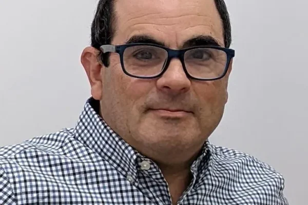 A man with short dark hair and glasses is wearing a checkered button-up shirt. He is looking directly at the camera with a neutral expression against a plain background
