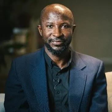 A man with a short beard and bald head is seated, wearing a dark blazer over a black shirt. He is looking directly at the camera with a neutral expression. The background is softly blurred, suggesting an indoor setting